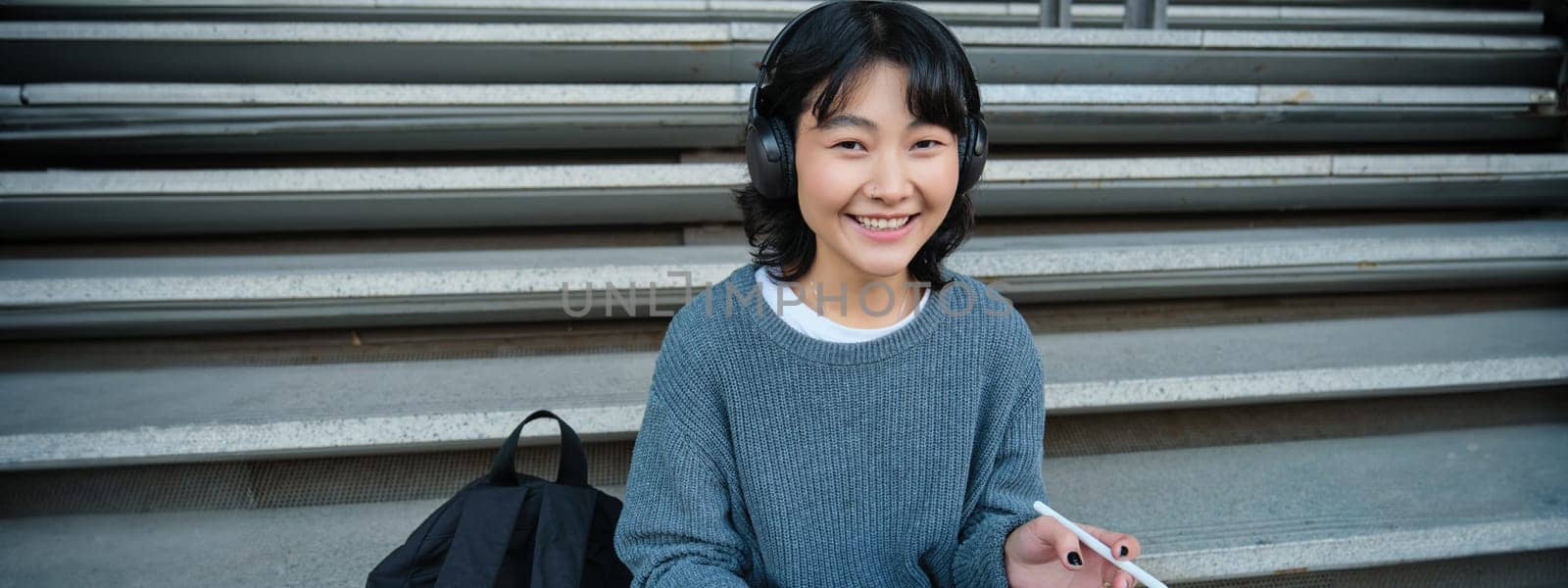 Young asian girl, student in headphones, works on remote, digital artist drawing on tablet with graphic pen, listening music in headphones and sitting on street staircase.