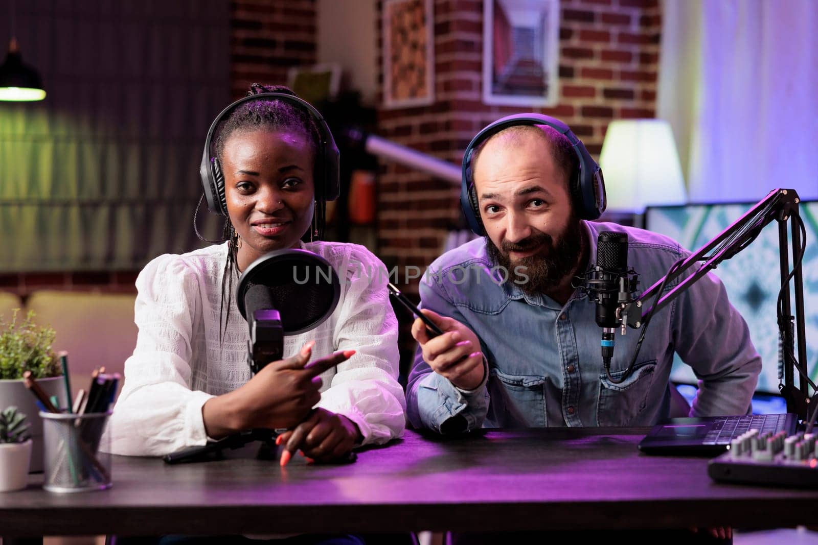 African american tech guru and colleague during online show in neon lights studio, shooting technology vlog using professional microphones. Vloggers presenting best smartphones on the market