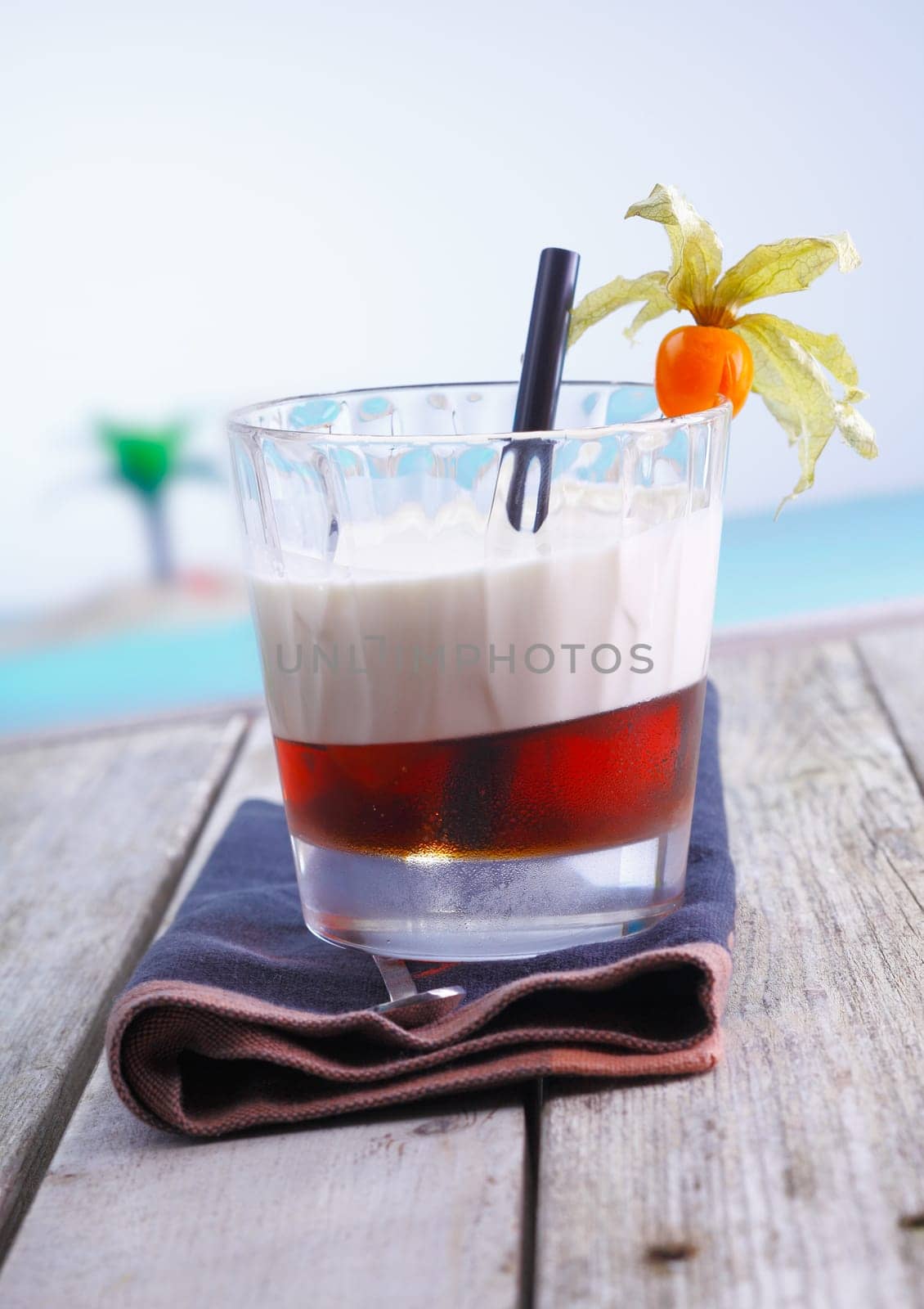 Layered alcoholic cocktail with gooseberry garnish served on a wooden deck by the sea