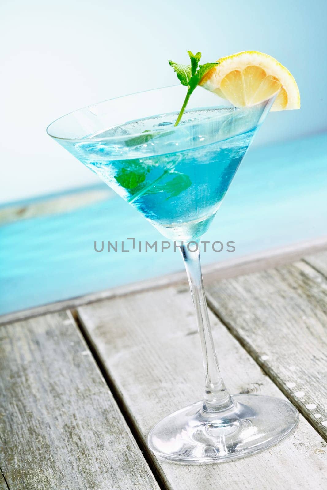 Refreshing classical blue curacao martini cocktail garnished with fresh lemon and served alongside the ocean on a tropical vacation