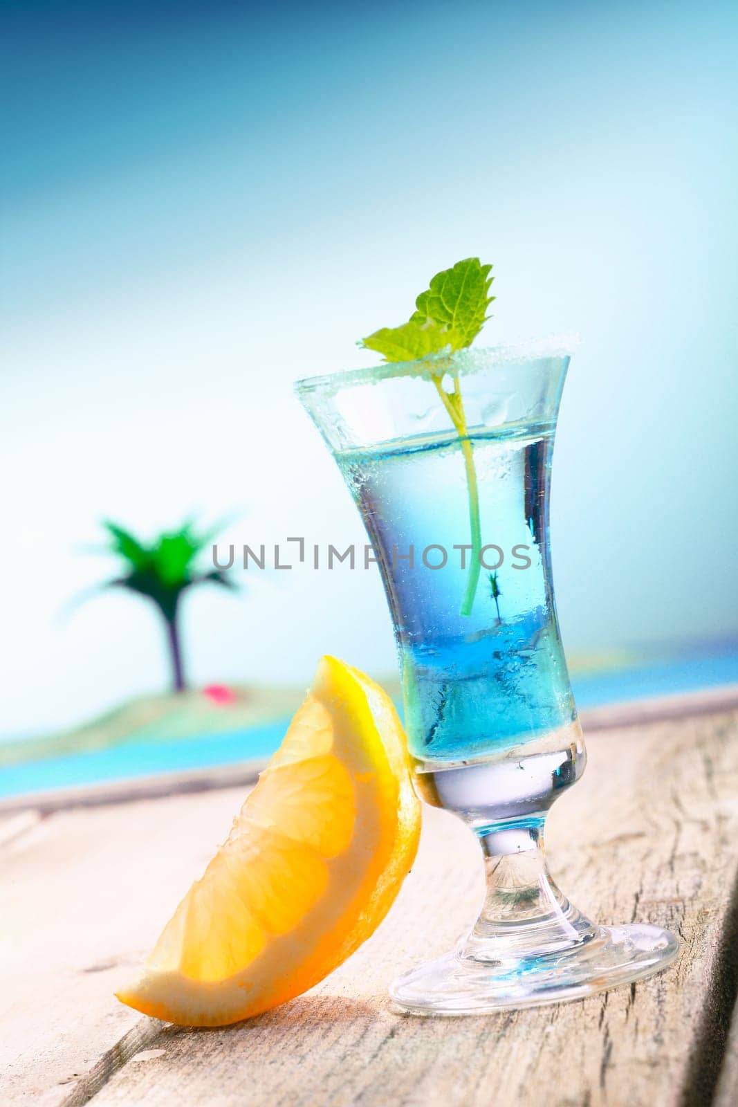 Blue Curacao Beach Cocktail with fresh lemon standing on a landing stage, with the pacific and a lagoon with a palm in the background. More Drinks in my portofolio.