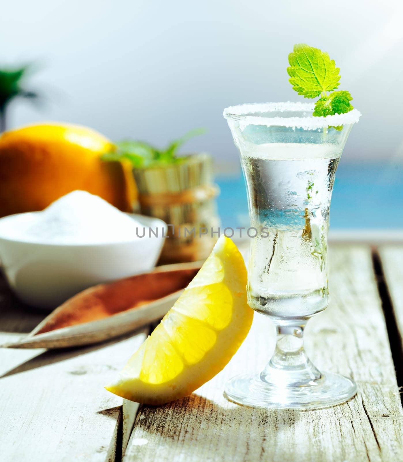 Chilled glass of vodka cocktail garnished with frosting and mint and served with lemon alongside a turquoise pool