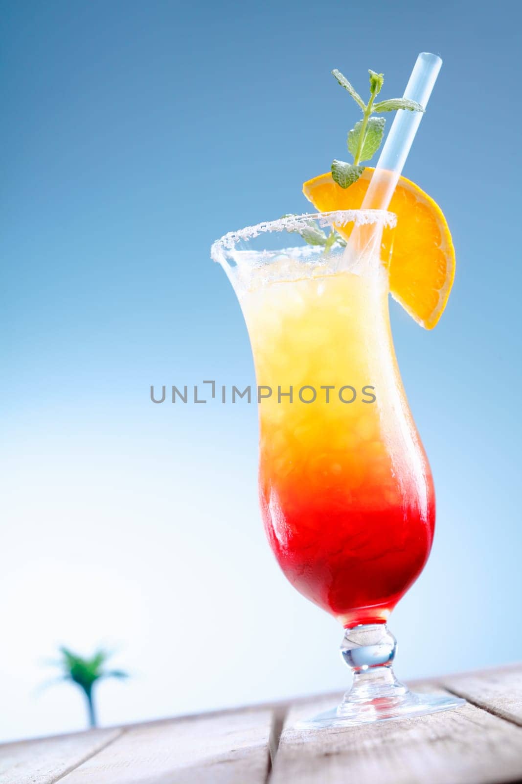 Refreshing cold glass of colourful Tequila Sunrise cocktail served on wooden decking against blue sky tilted at an angle