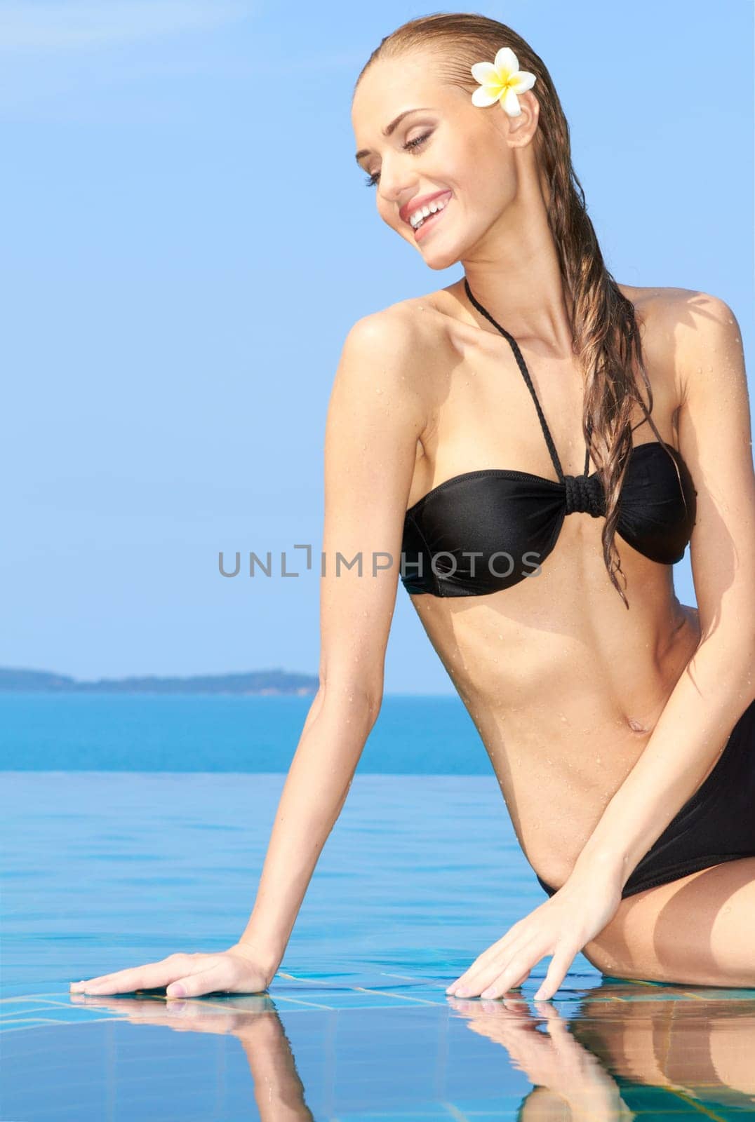 Smiling woman with wet hair sitting on the tiled edge of the pool reflected in the water below
