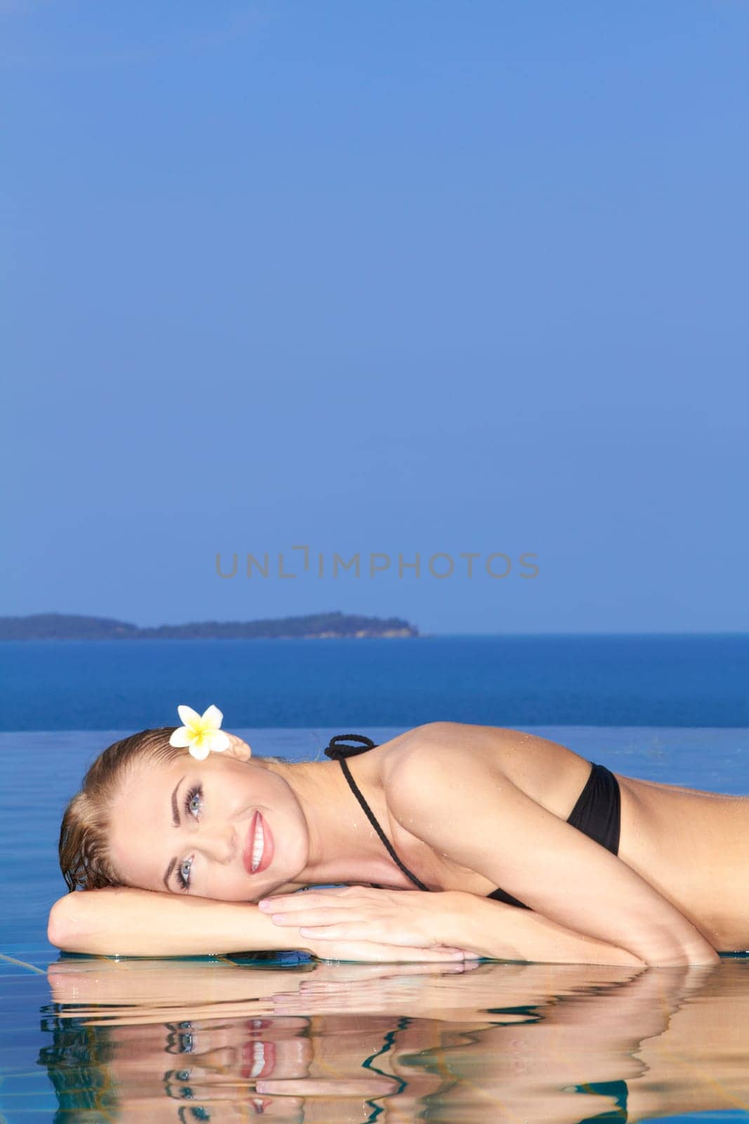 Smiling woman with wet hair laying with her arms on the tiled edge of the pool reflected in the water below