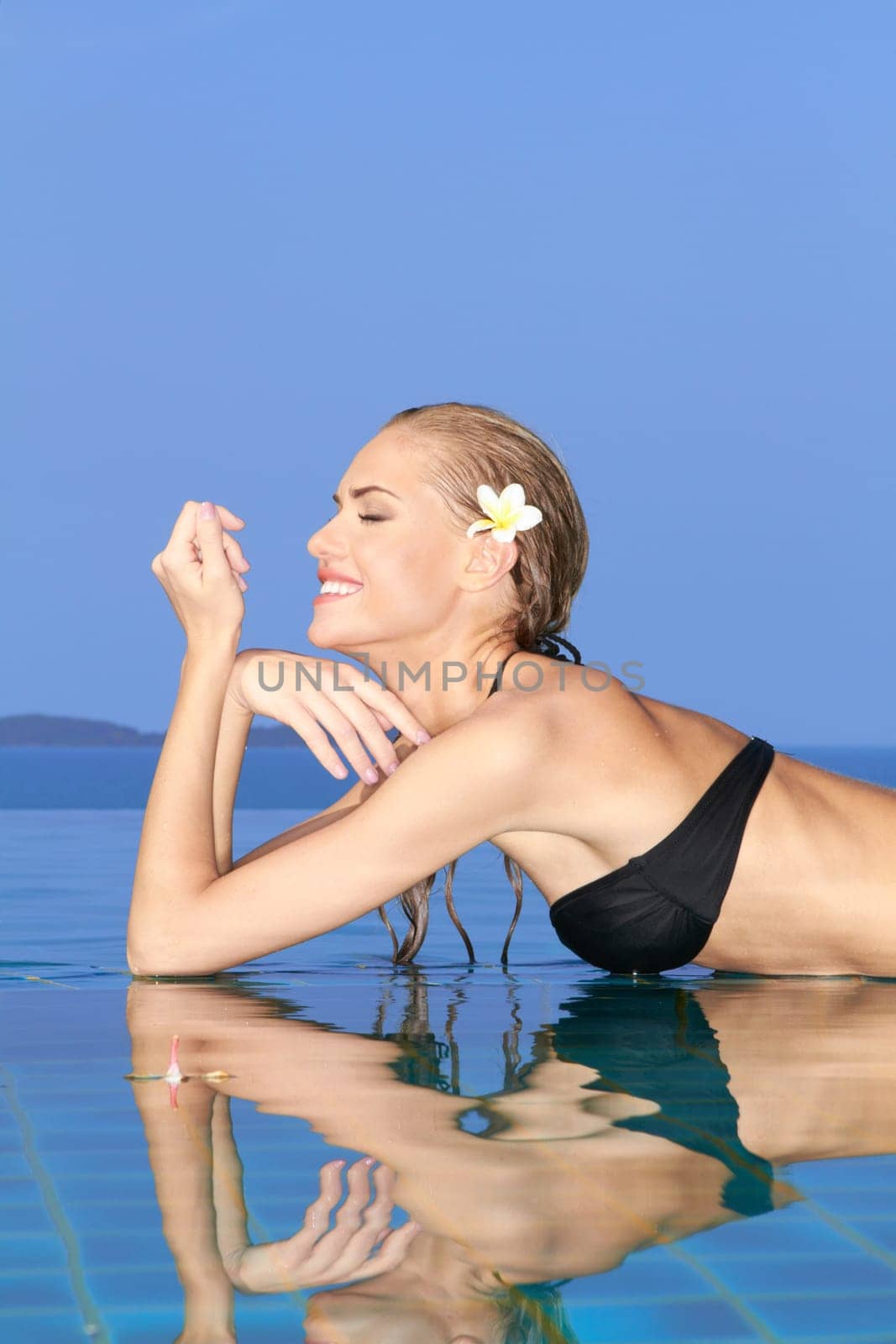 Smiling woman with wet hair laying with her arms on the tiled edge of the pool reflected in the water below