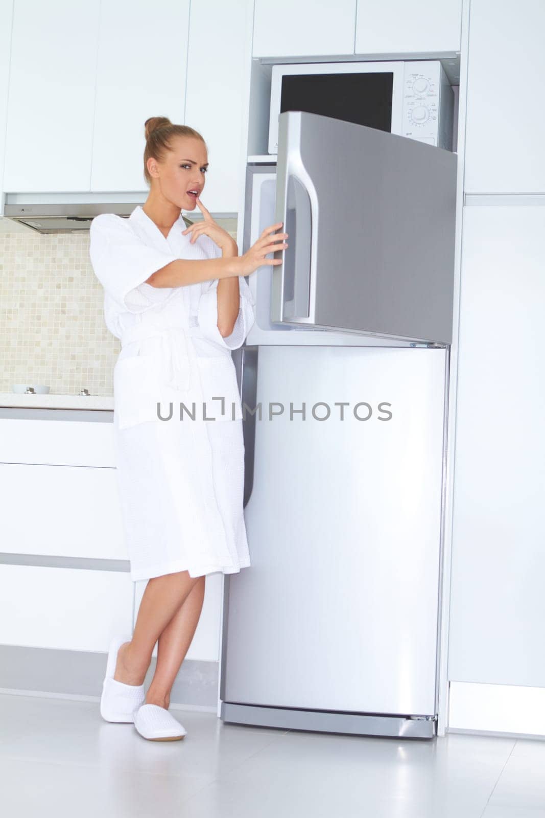 Vivacious woman in a white bathrobe and slippers standing laughing as she opens the freezer unit of her refrigerator