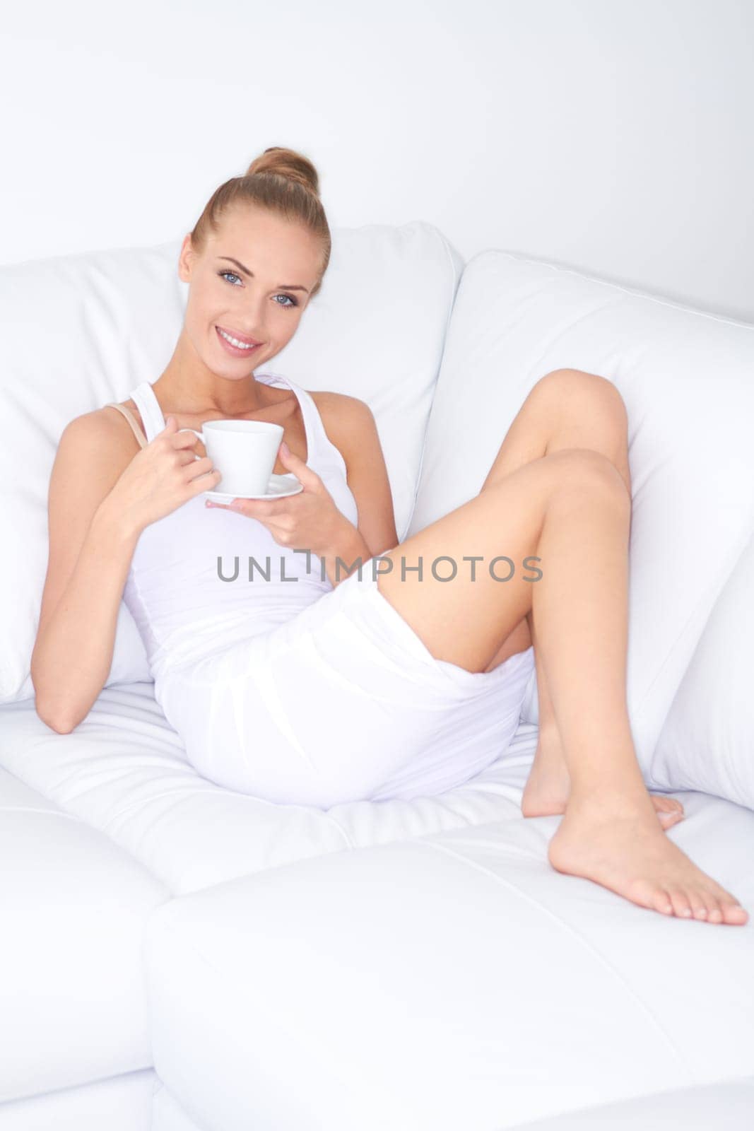 Beautiful elegant woman barefoot in a white dress reclining on a sofa drinking coffee