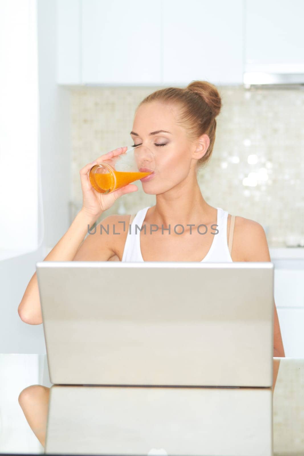 Healthy happy woman drinking from glass of fresh orange juice seated at a table using her laptop