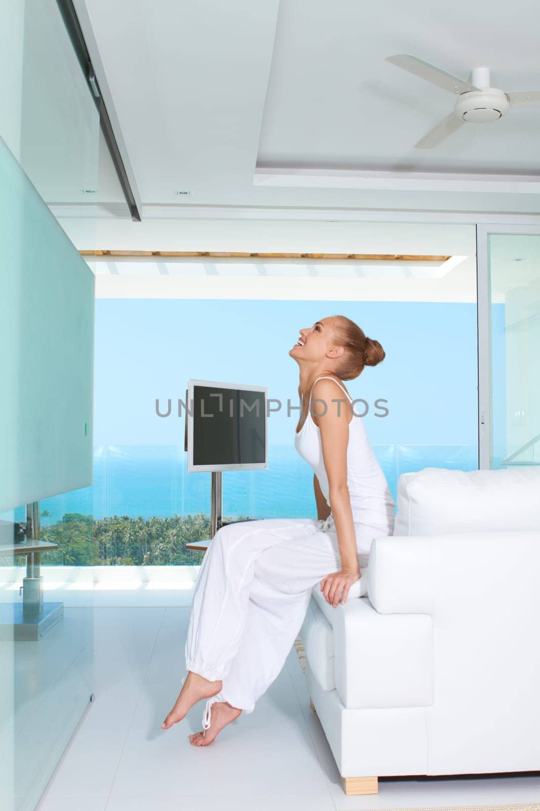 Graceful woman in white baggy slacks sitting barefoot on the edge of a white sofa in a modern glass fronted living-room with ocean view