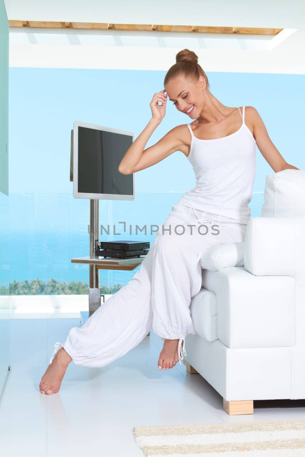 Graceful woman in white baggy slacks sitting barefoot on the edge of a white sofa in a modern glass fronted living-room with ocean view