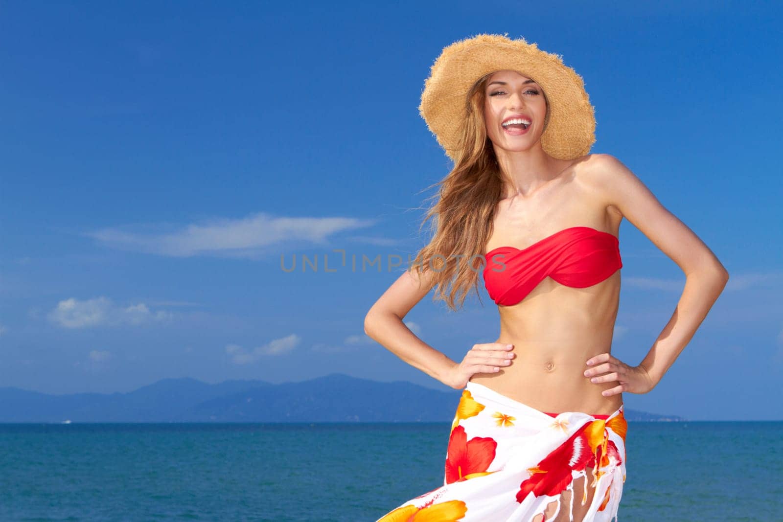 High-spirited beautiful woman laughing in merriment as she poses in front of the ocean in her bikini and colourful sarong