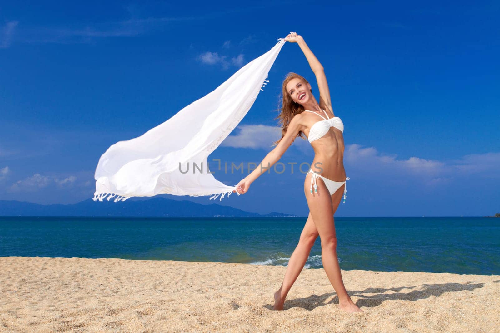 Smiling woman in white bikini holding white scarf against blue sky ,  on beautiful sandy beach