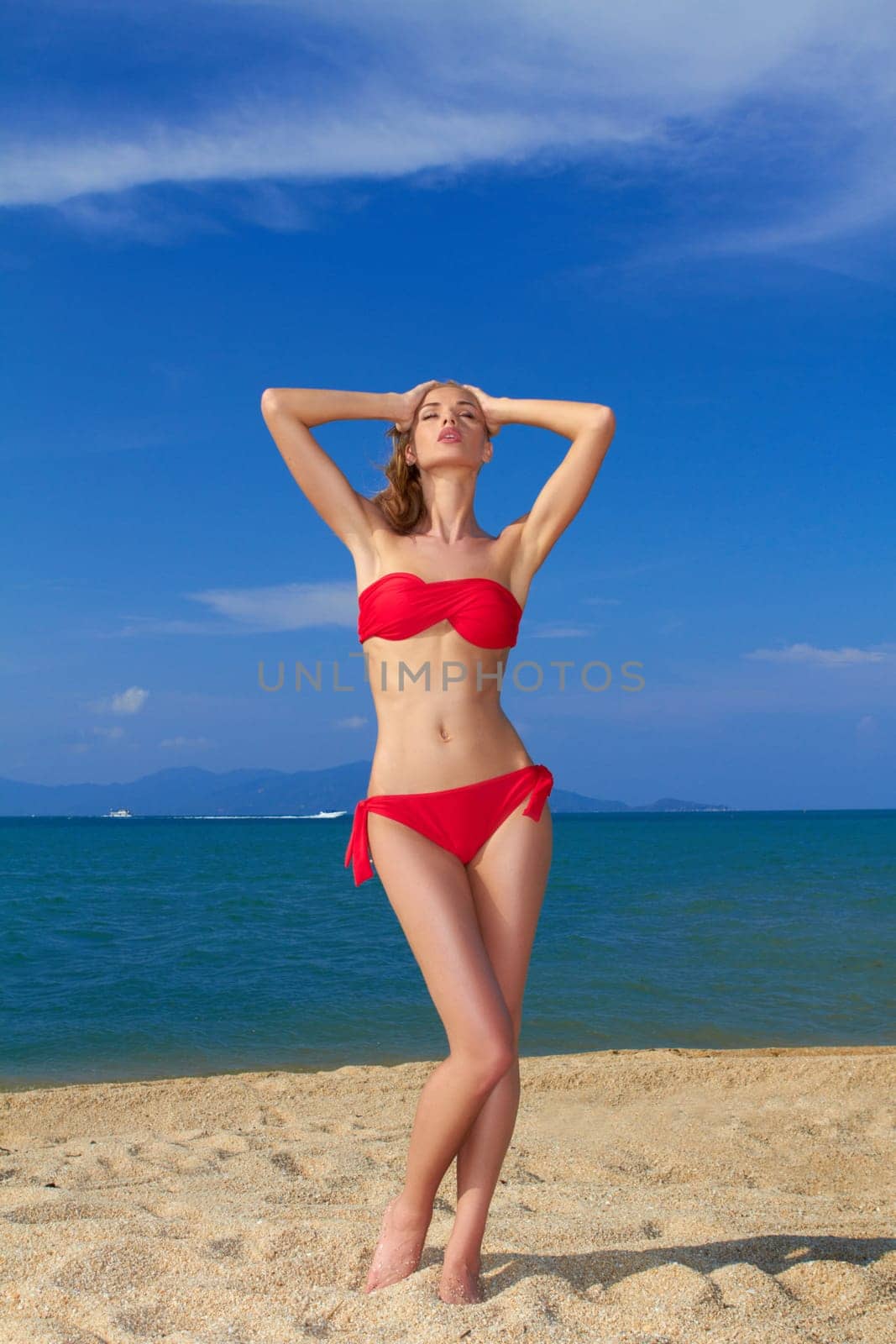 Sexy woman in red bikini posing on a tropical beach with ocean backdrop with her arms raised above her head
