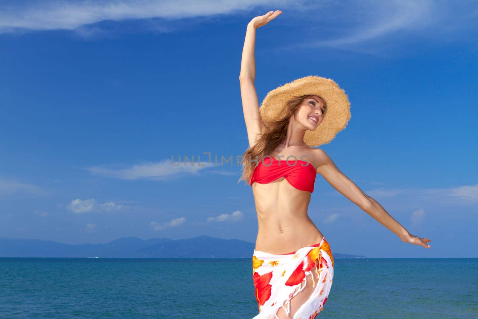 Beautiful lively woman full of the joys of life twirling with her arms in the air in a colourful bikini and sarong against an ocean backdrop