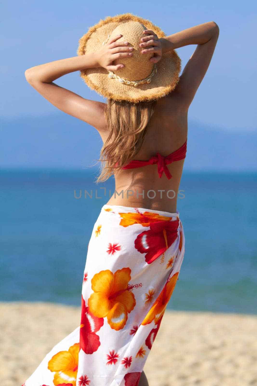 Woman in a colourful tropical sarong and straw hat standing on a beach facing away from the camera out over the ocean with her arms raised