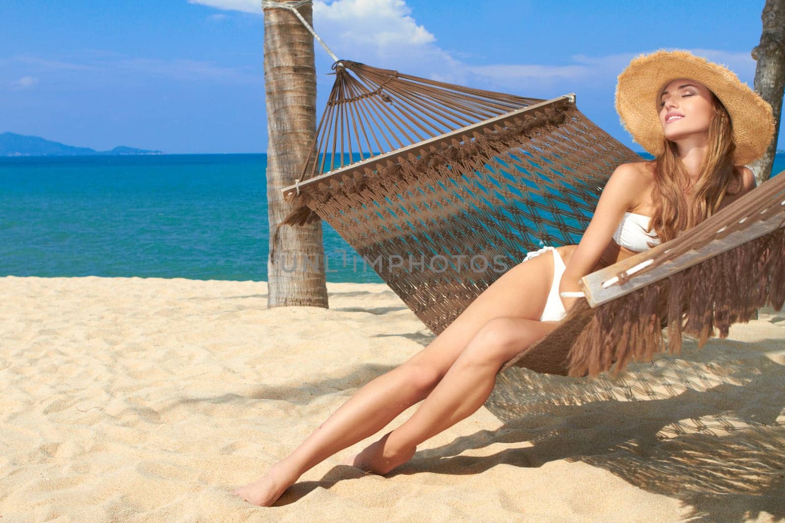 Elegant woman in a bikini reclining in a hammock strung between palm trees on the beach at a tropical resort
