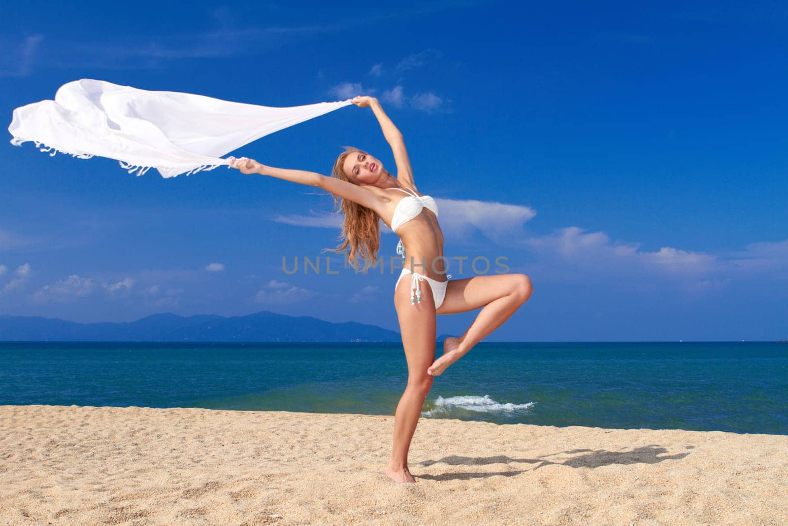 Bikini clad blonde beauty in a dancers pose on soft white sand with the ocean in the background