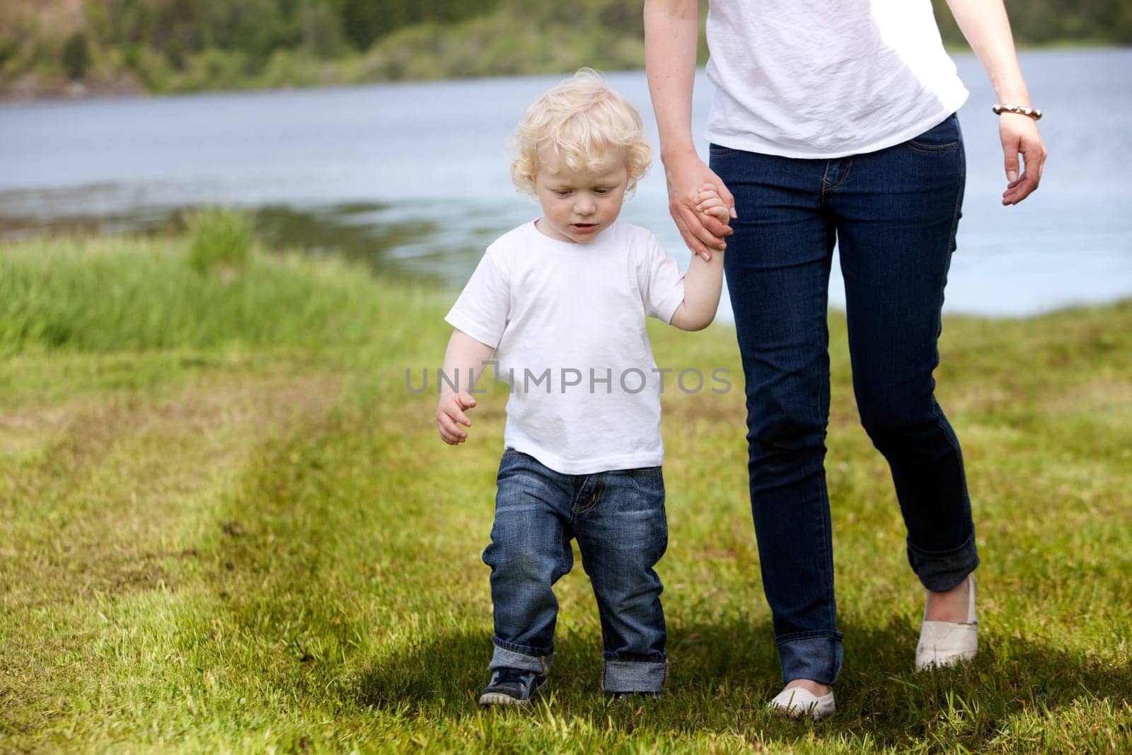 A young child walking in grass with his mother near a lake