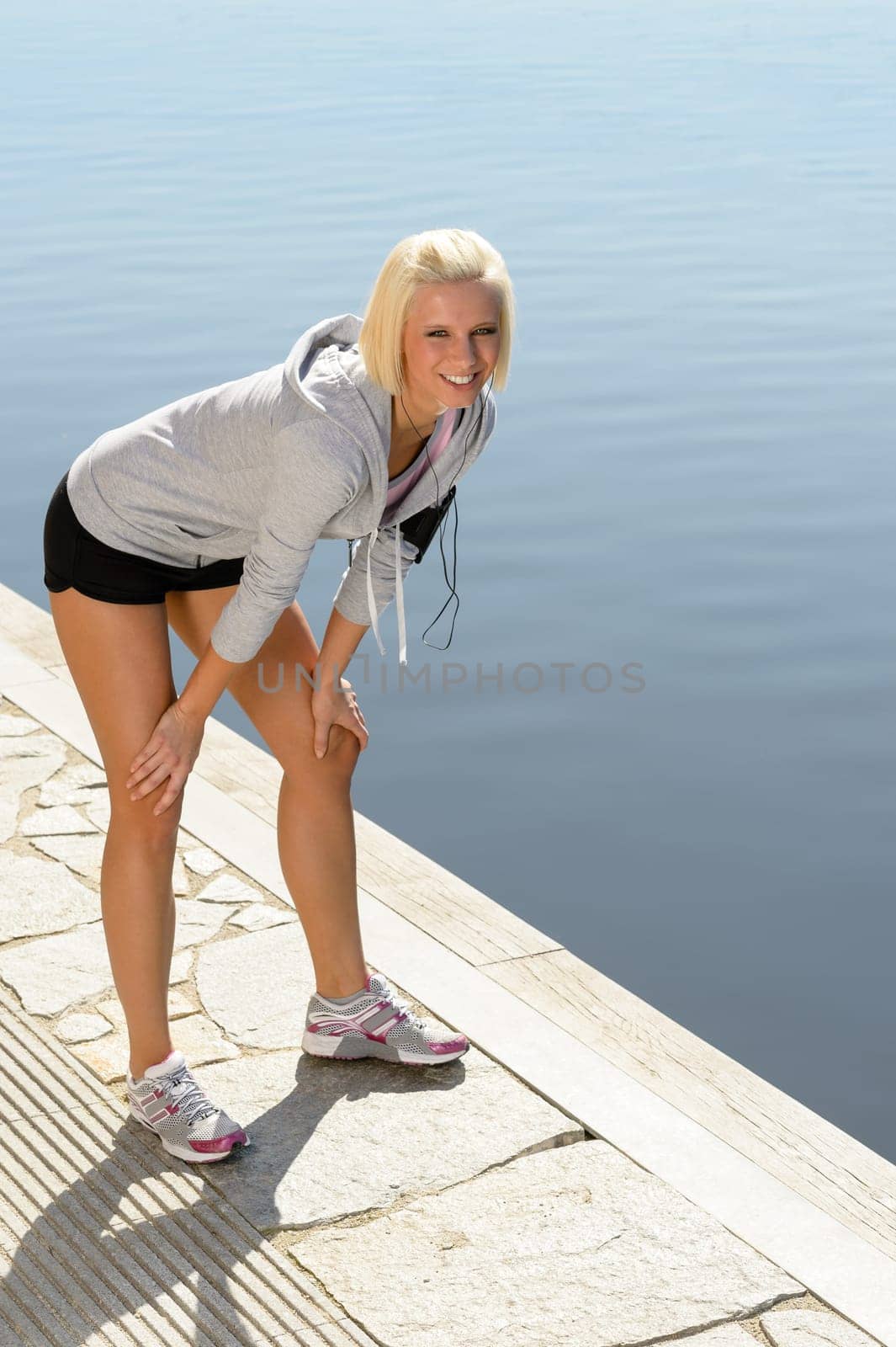 Sport blond woman stretching legs on a pier sunny day