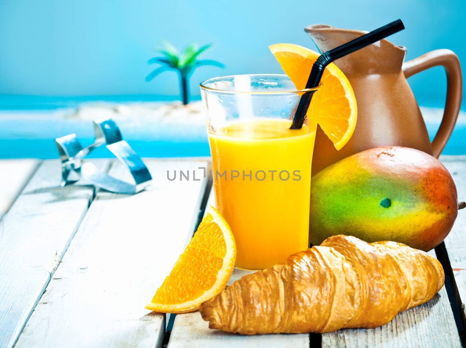 Tropical breakfast still life with a healthy mango and orange smoothie and a fresh ripe mango with a golden croissant against a blue ocean backdrop