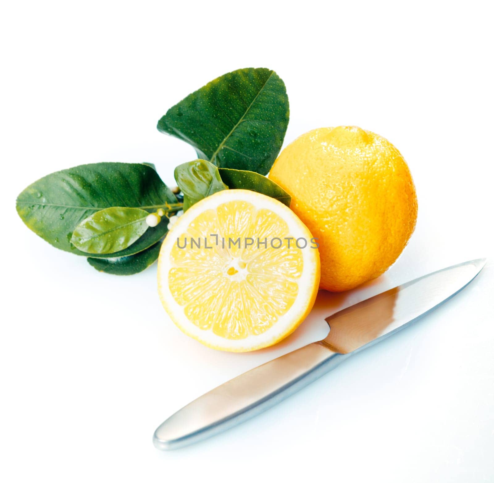 Halved and whole fresh lemons with leaves and a knife on a white studio background