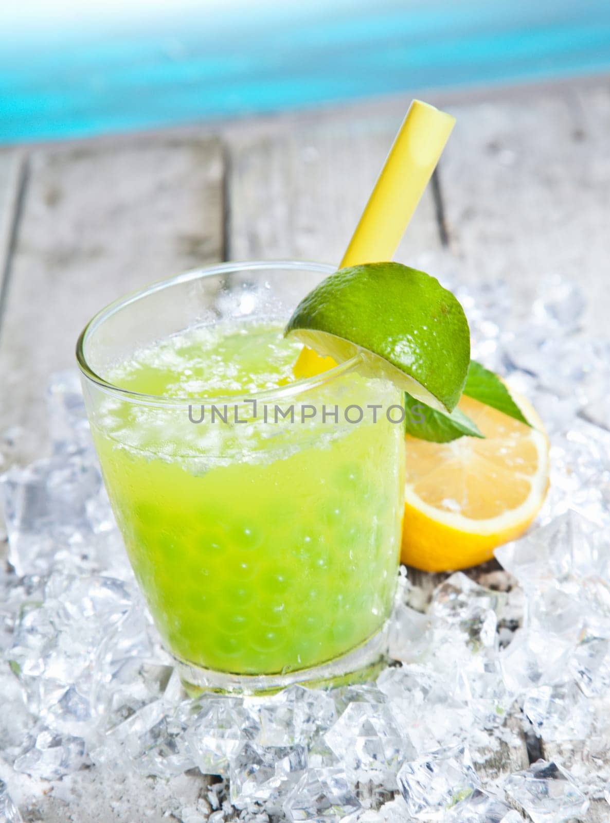 Glass of tangy refreshing lime boba tea cocktail standing on a bed of crushed ice on a wooden deck alongside blue water