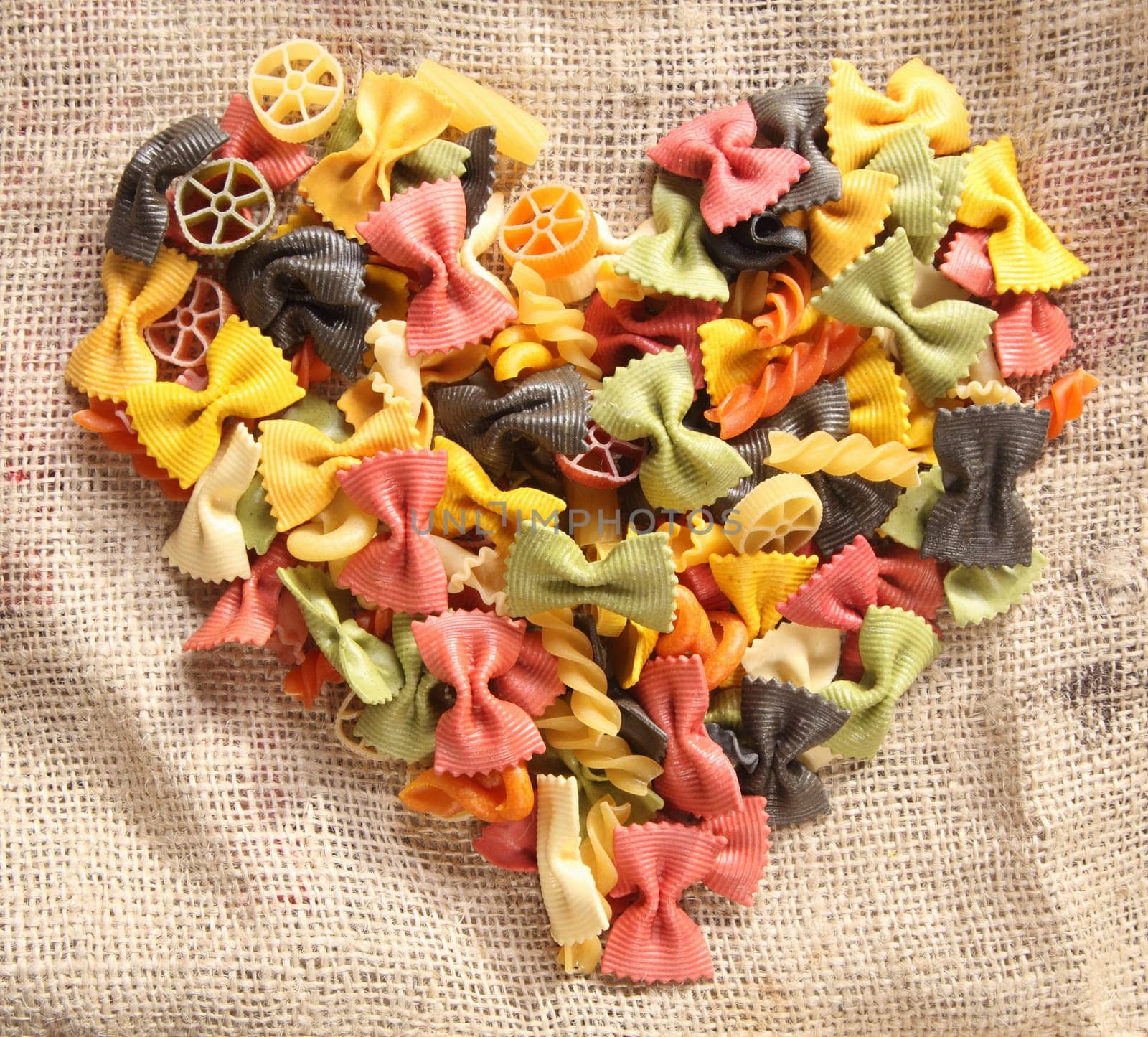 Colorful Bowtie Pasta Heart. Heart-shaped arrangement of colourfaul bowtie pastas, or farfalle, on rustic hessian material, concpetual of love and romance.