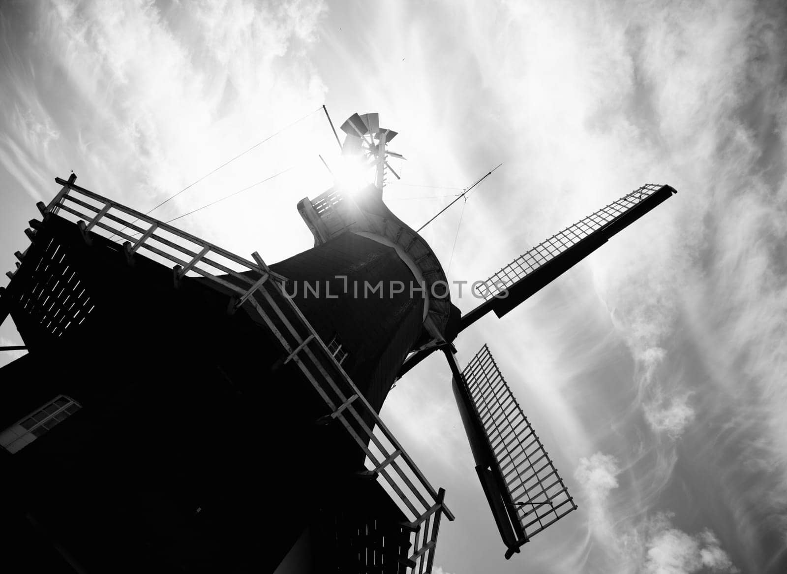 Monochrome image of a tilted windmill with it sails and balcony silhouetted against a cloudy sky by a sun flare over the roof.