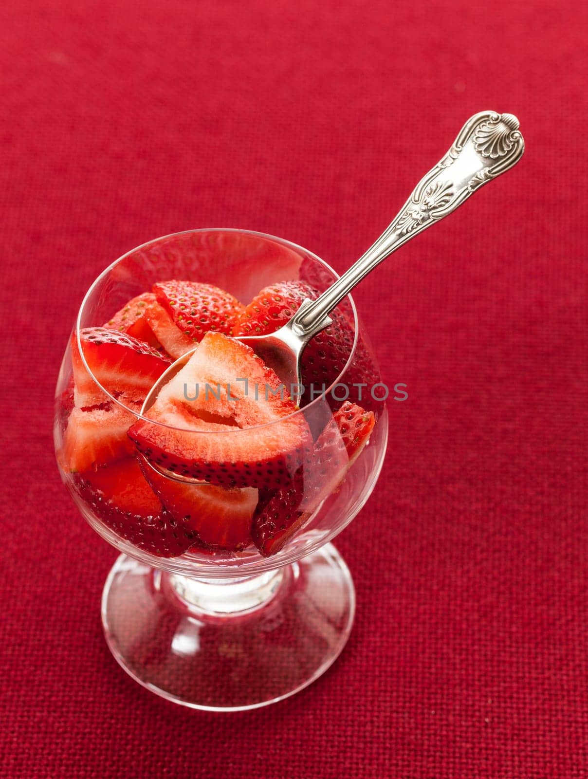 Glass dish with freshly sliced strawberry fruits against a red tablecloth with silver spoon