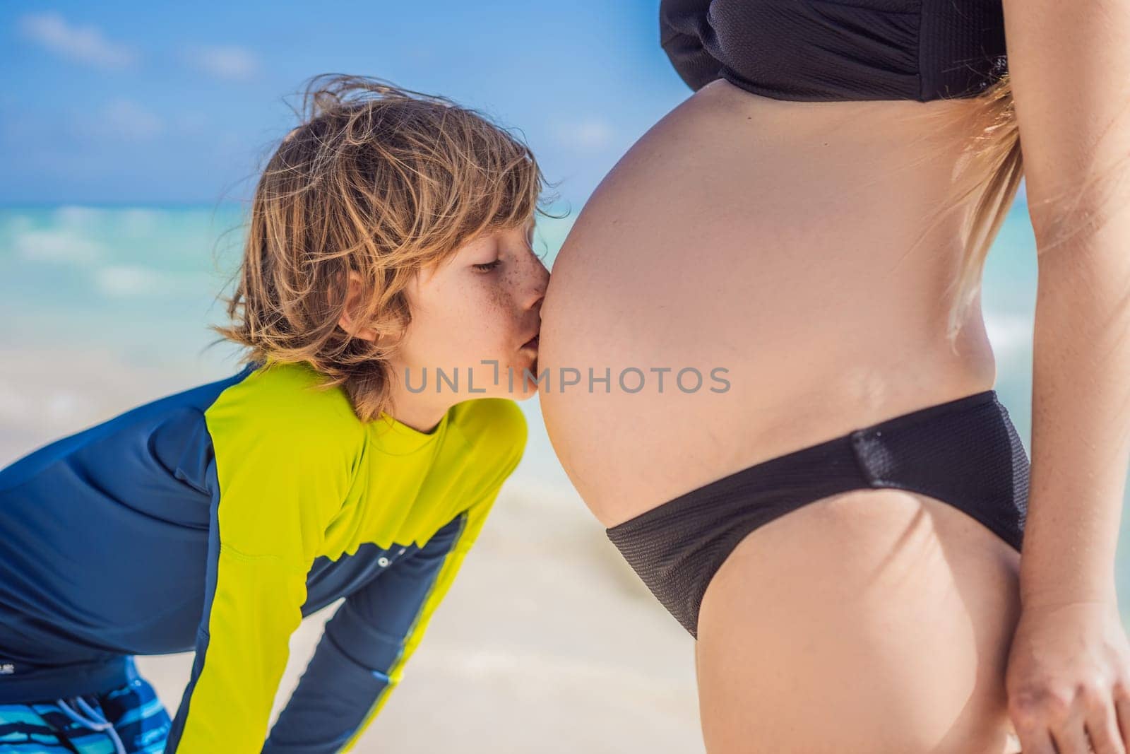 A radiant pregnant mother and her excited son share a tender moment on a serene, snow-white beach, celebrating family love amidst nature's beauty by galitskaya