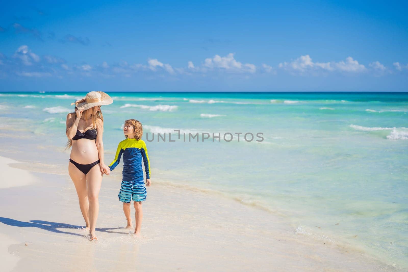 A radiant pregnant mother and her excited son share a tender moment on a serene, snow-white beach, celebrating family love amidst nature's beauty.