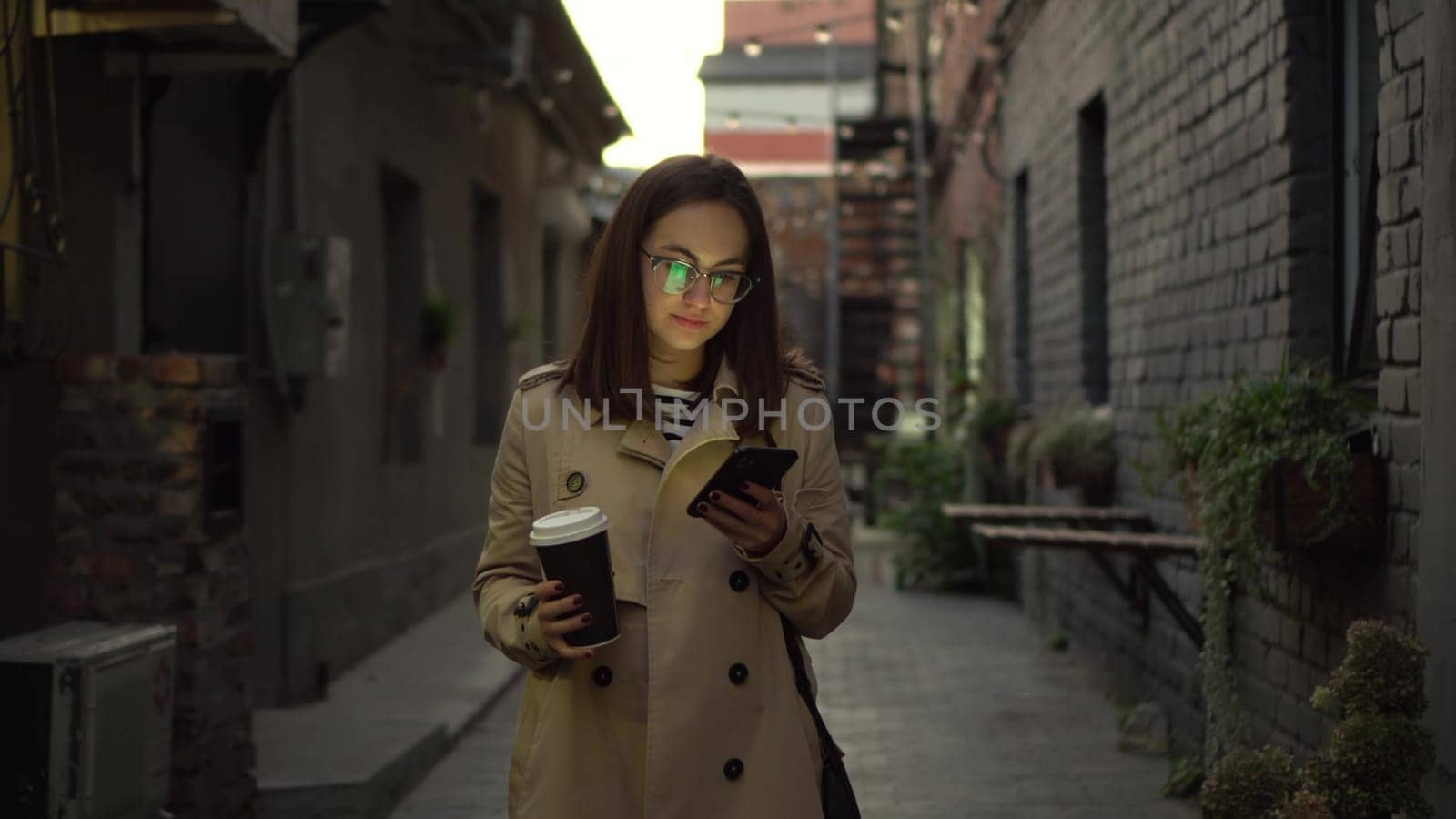 A young woman walks along a narrow street late at night and chats on her smartphone. A girl with glasses walks with a phone in her hands and drinks coffee. 4k