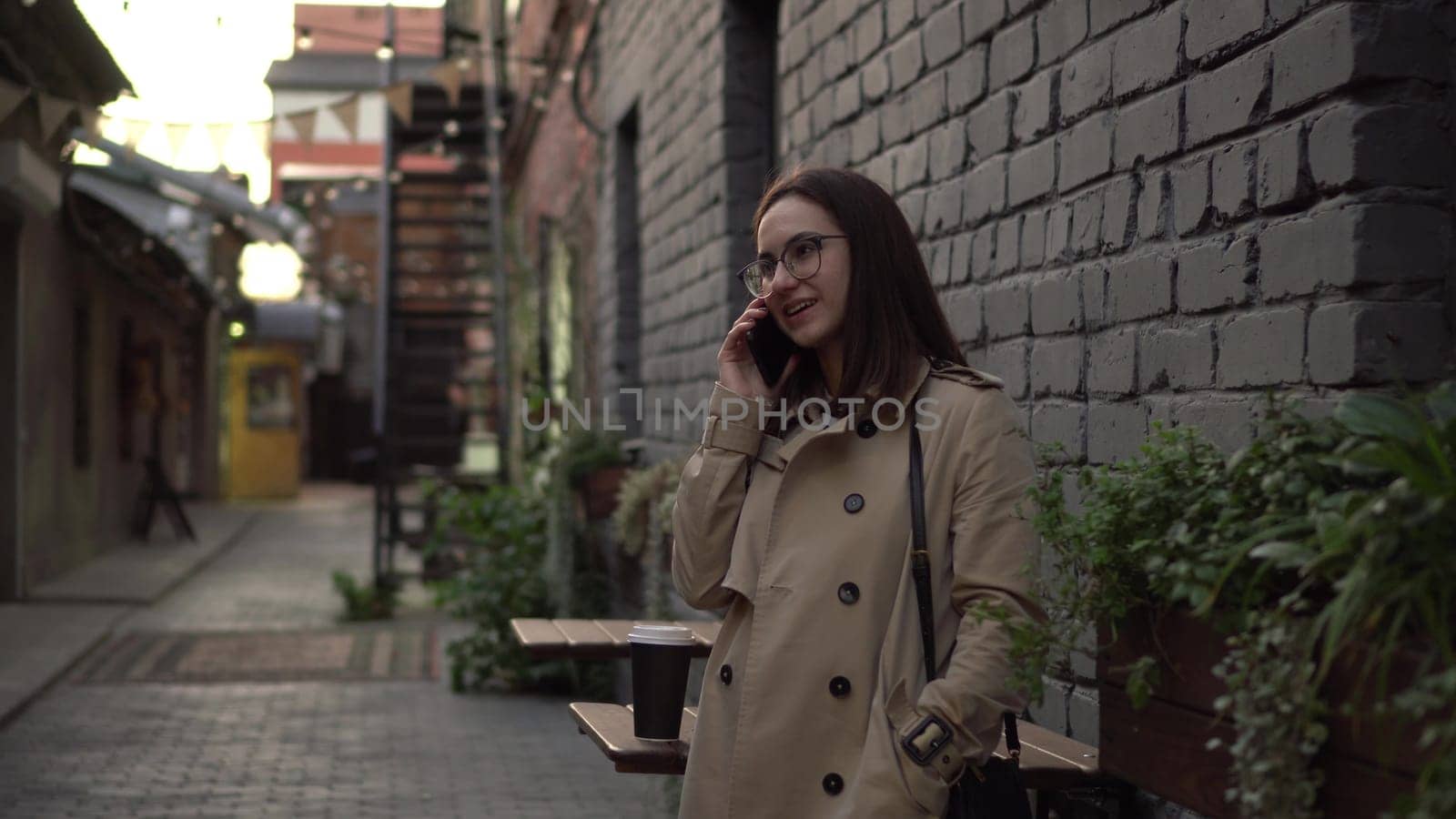 A young woman talks on the phone while standing against a wall on a narrow street. A girl with glasses and a coat speaks on the phone. 4k