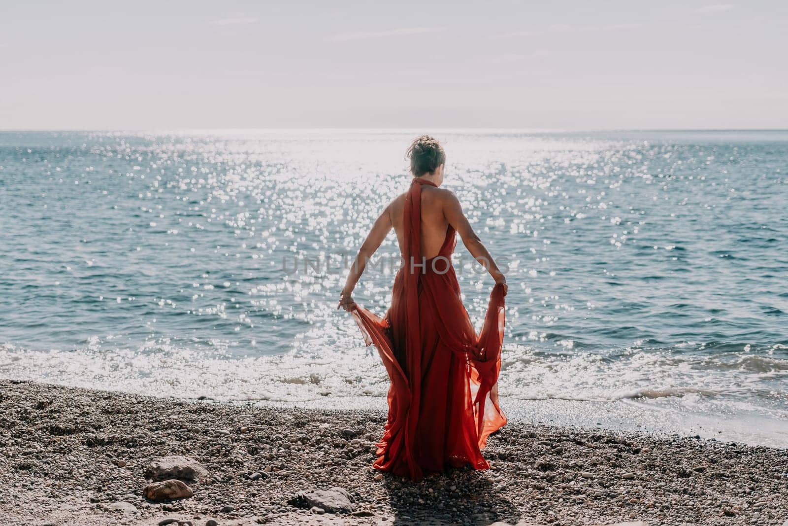 Side view a Young beautiful sensual woman in a red long dress posing on a rock high above the sea during sunrise. Girl on the nature on blue sky background. Fashion photo.