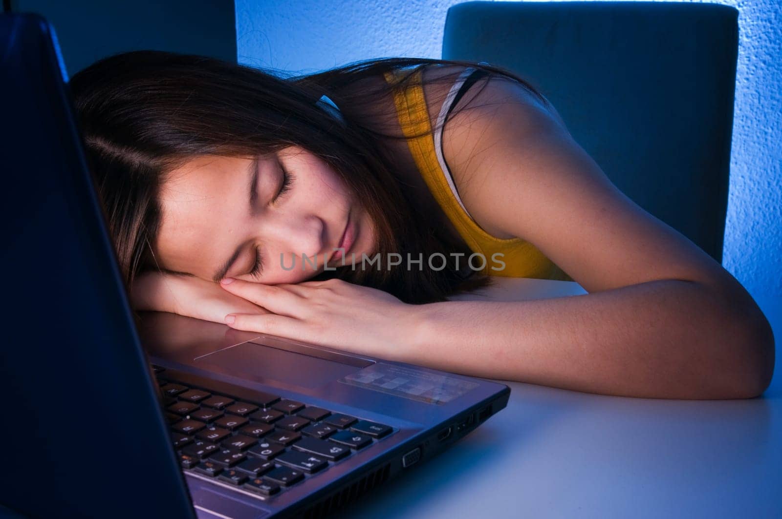 Head and shoulders shot of a young black-haired woman with her head resting on her hands in front of the computer keyboard sleeping in wide-angle and sideways view at table height
