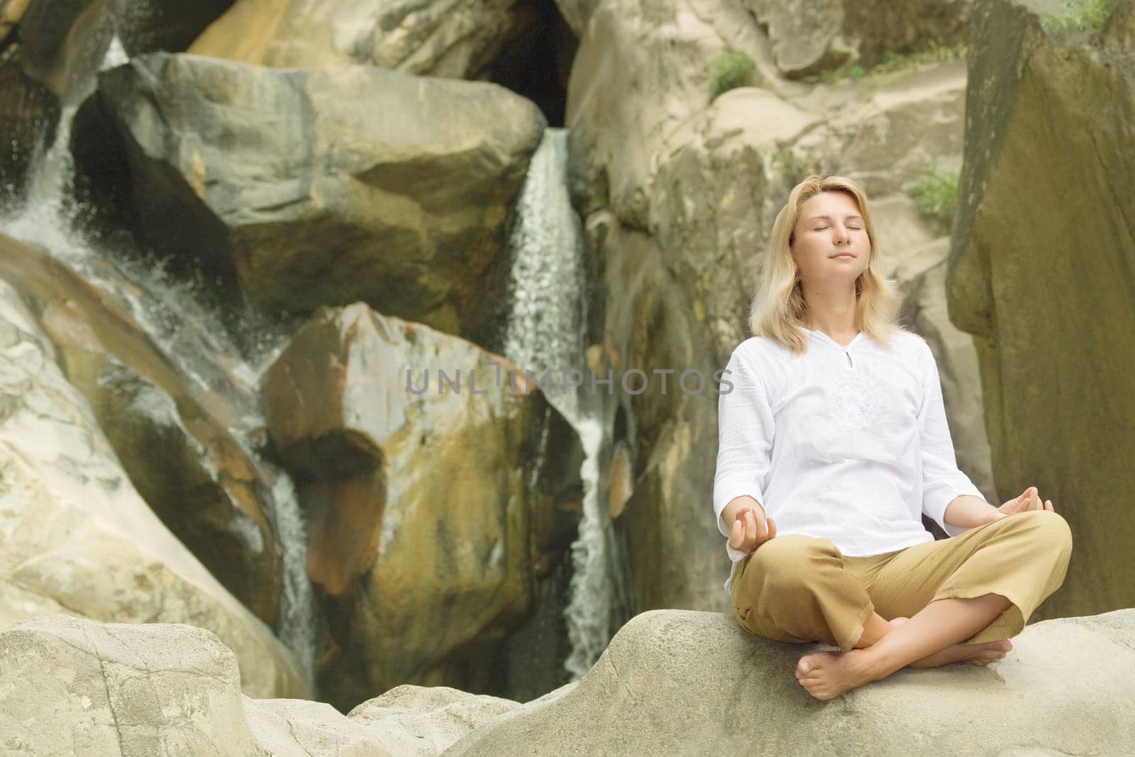 Young woman doing yoga exercises over waterfall background.