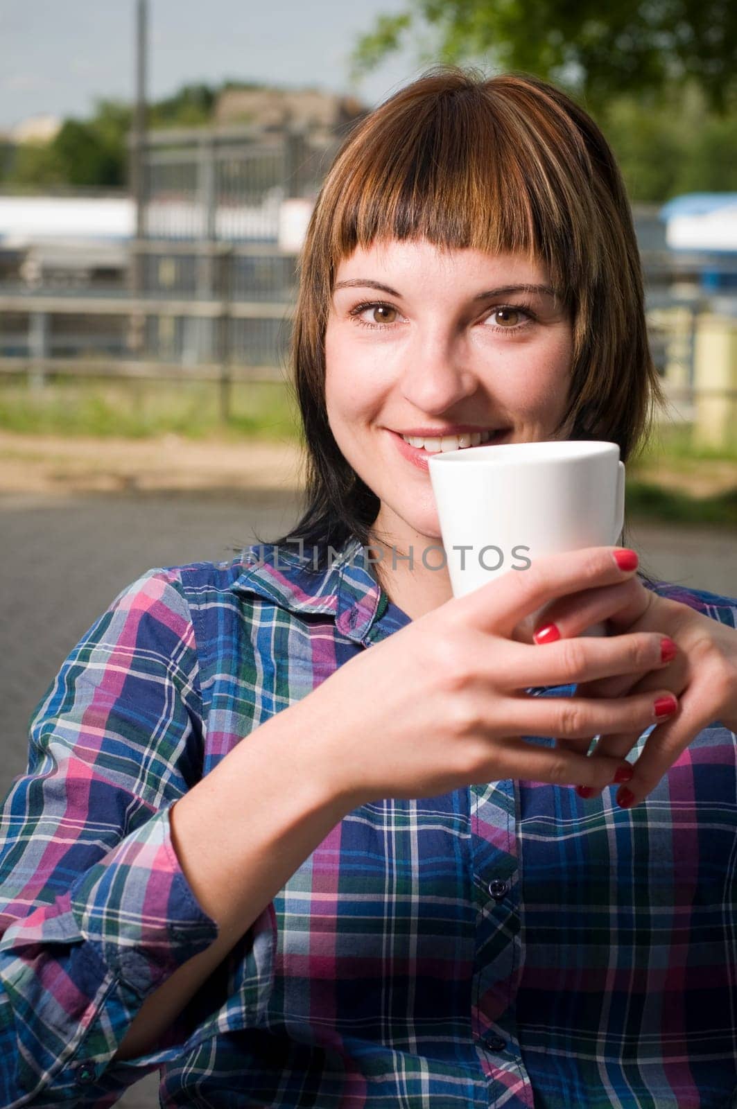 Head and shoulders shot of a young brown-haired woman sitting at a table in a street café and holding a white cup