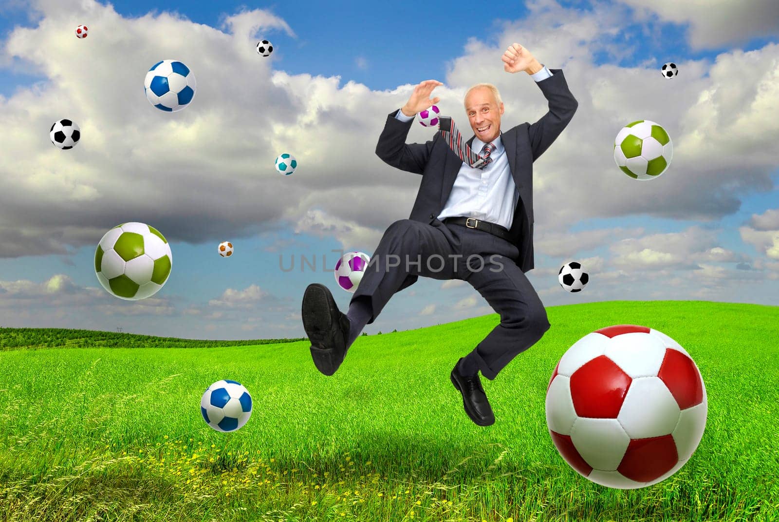 Happy mature businessman jumping outdoors in a green field with soccer balls all around