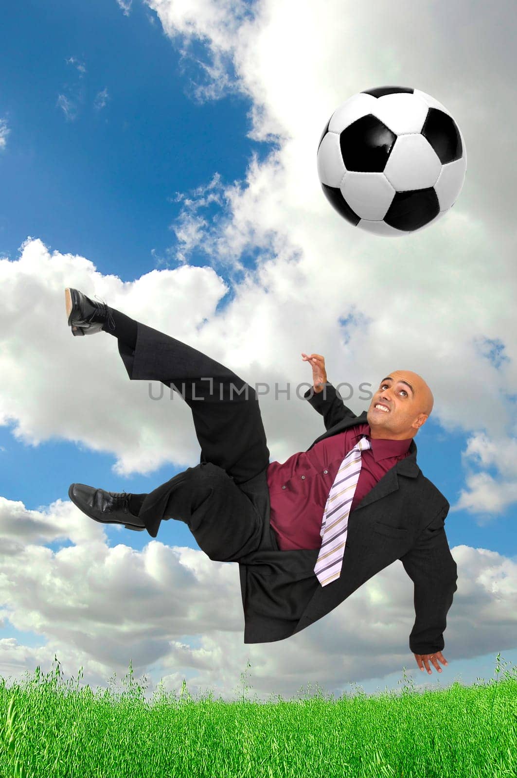 Businessman in a acrobatic pose kicking a soccer ball in a green field