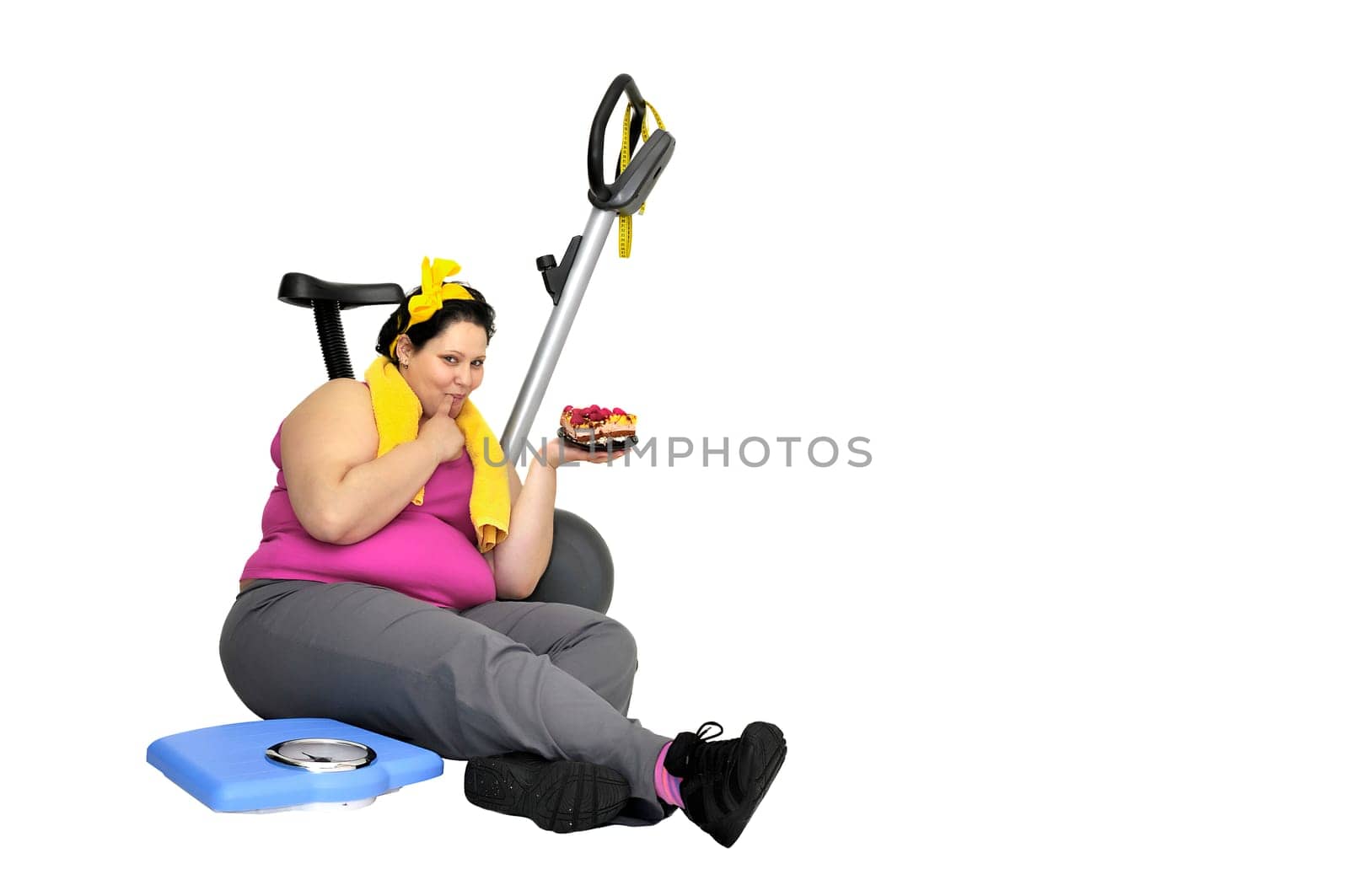 Large girl with fitness bicycle and a scale, holding a cake isolated in white