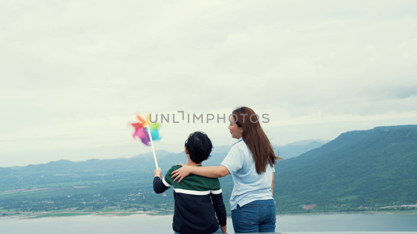 A progressive woman and her son are on vacation, enjoying the natural beauty of a lake at the bottom of a hill while the boy carries a toy windmill.