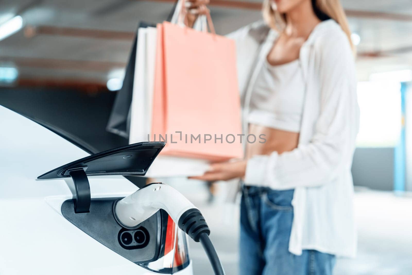 Young woman travel with EV electric car to shopping center parking lot charging in downtown city showing urban sustainability lifestyle by green clean rechargeable energy of electric vehicle innards