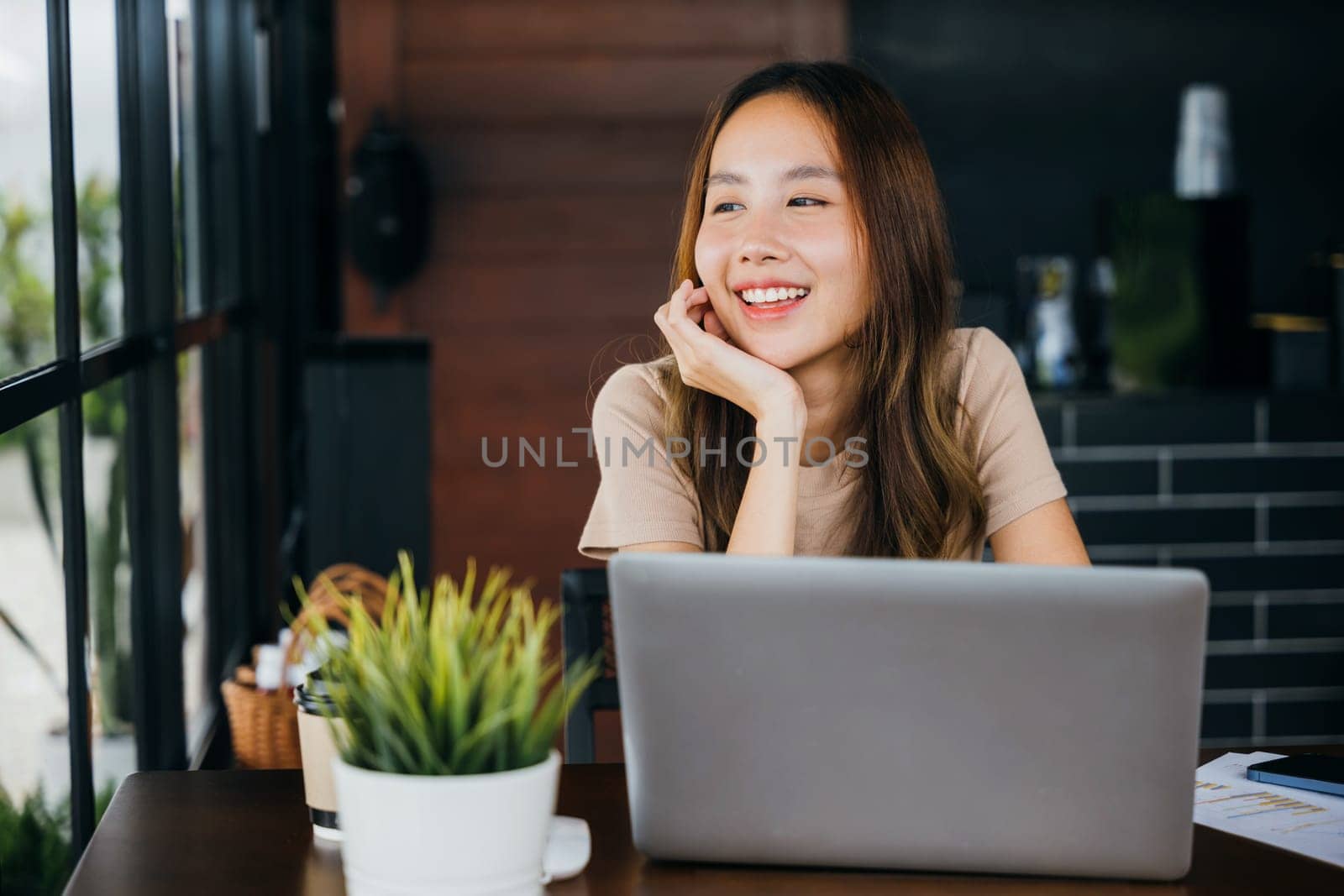 Happy Asian young businesswoman sitting alone at cafe desk with laptop computer she looking out of window, charming woman working outside and smiling in coffee shop freelancer thinking about new ideas