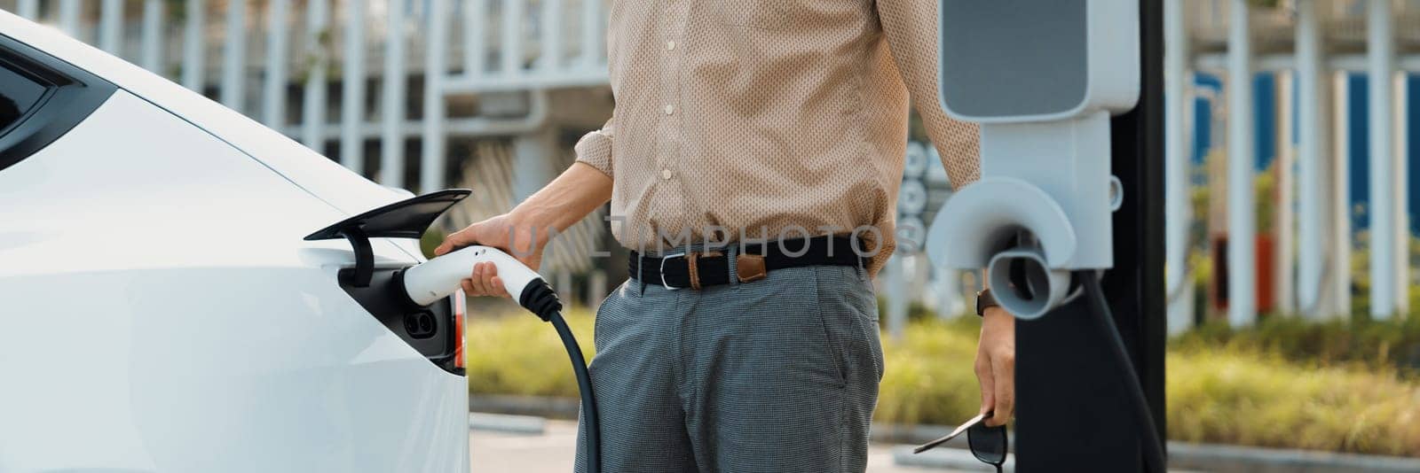 Young man recharge electric car's battery from charging station in city commercial parking lot. Rechargeable EV car for sustainable environmental friendly urban travel. Panorama Expedient