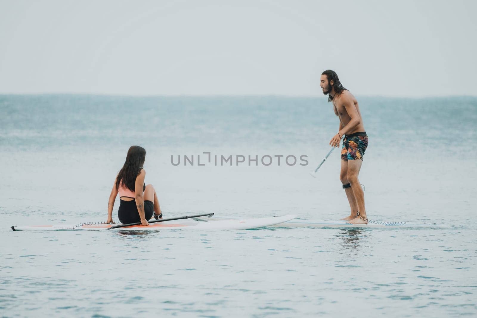 Sea woman and man on sup. Silhouette of happy young woman and man, surfing on SUP board, confident paddling through water surface. Idyllic sunset. Active lifestyle at sea or river