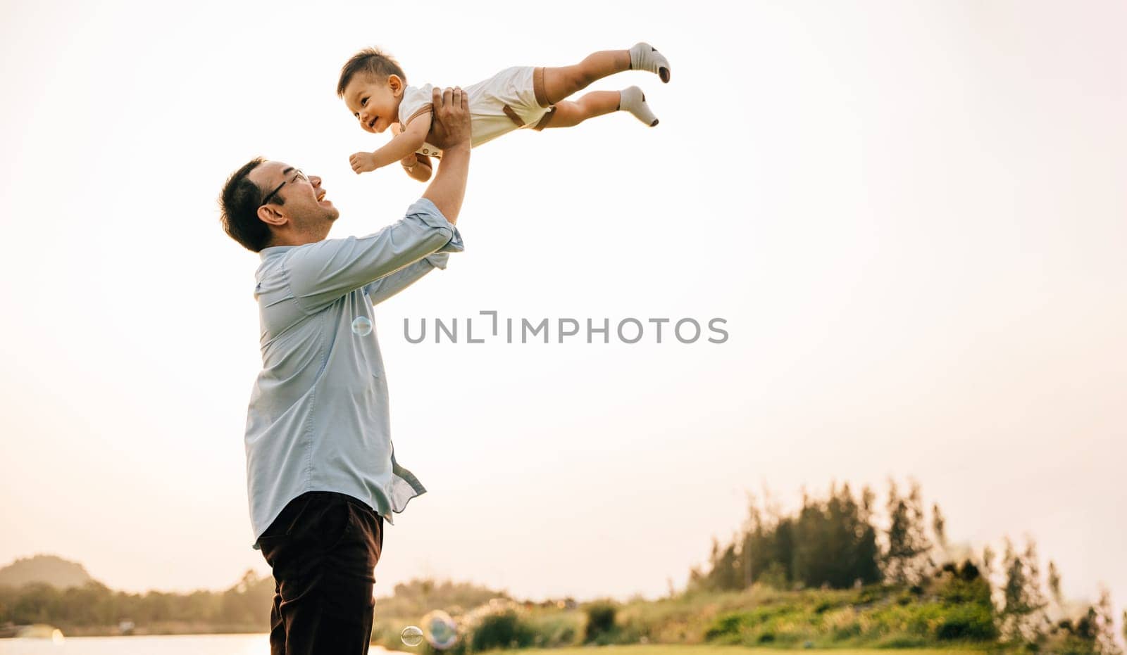 Playful family moment, father holds happy little boy up high, throwing up in sky on his birthday. cheerful child enjoys moment of freedom and joy, while the loving dad captures precious memory forever