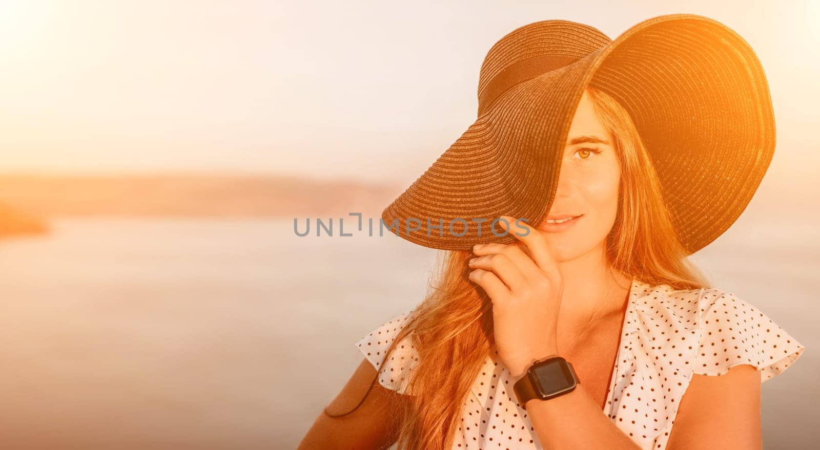 Portrait of happy young woman wearing summer black hat with large brim at beach on sunset. Closeup face of attractive girl with black straw hat. Happy young woman smiling and looking at camera at sea