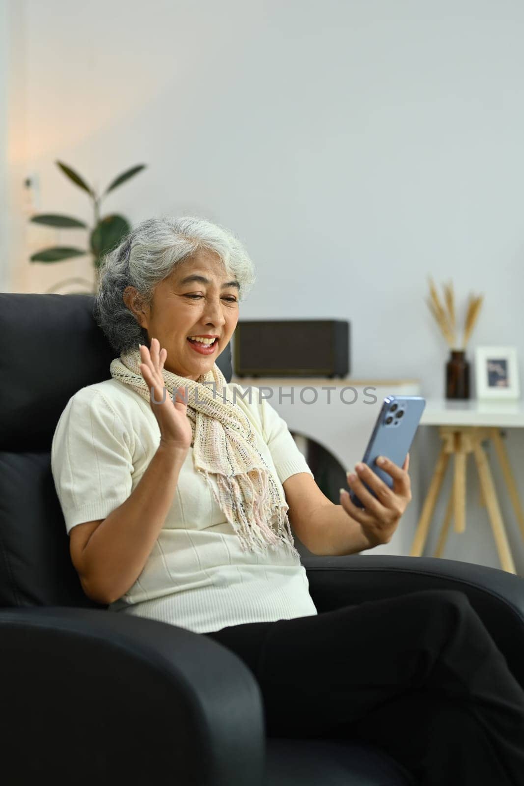 Beautiful gray haired mature woman waving hand while talking online on smartphone by prathanchorruangsak