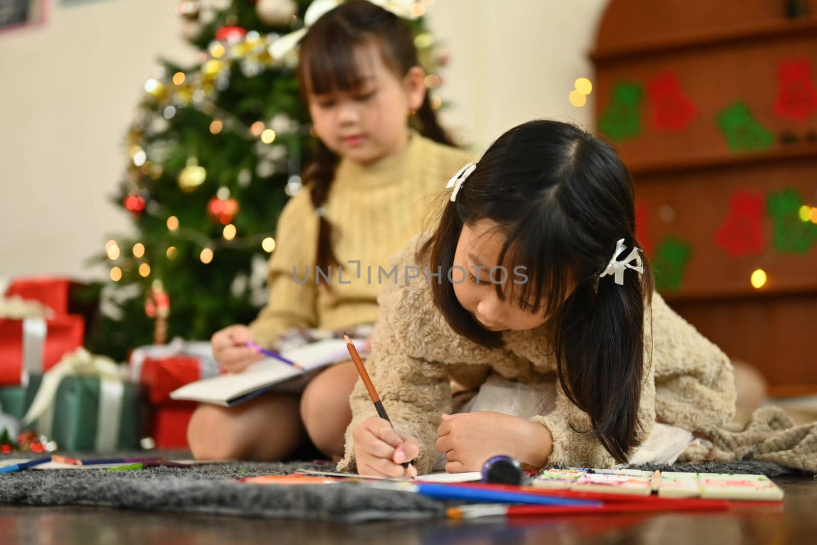 Adorable little girl making greeting card for New Year and Christmas while lying on carpet in decorated living room.